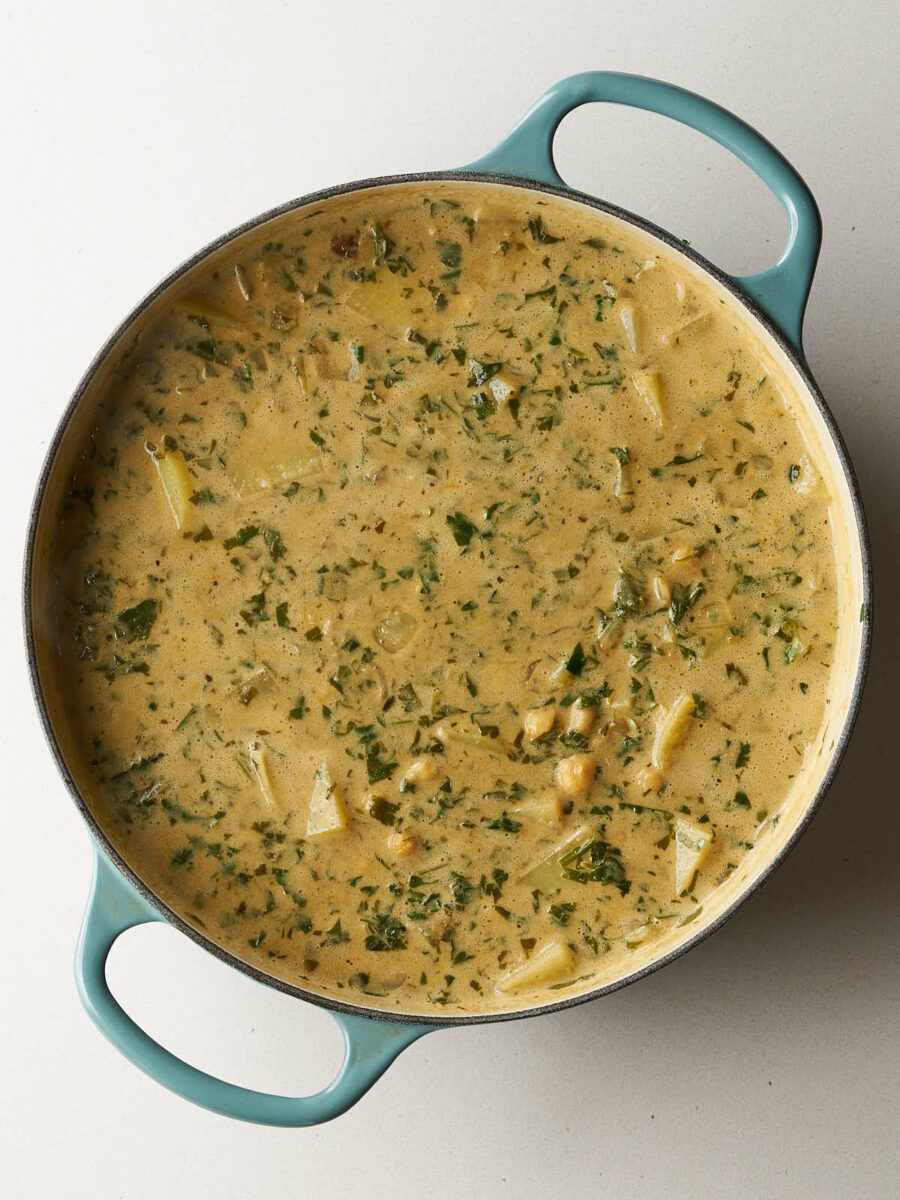 A creamy stew base with chunks of potatoes, chickpeas, and flecks of green herbs in a light blue dutch oven.