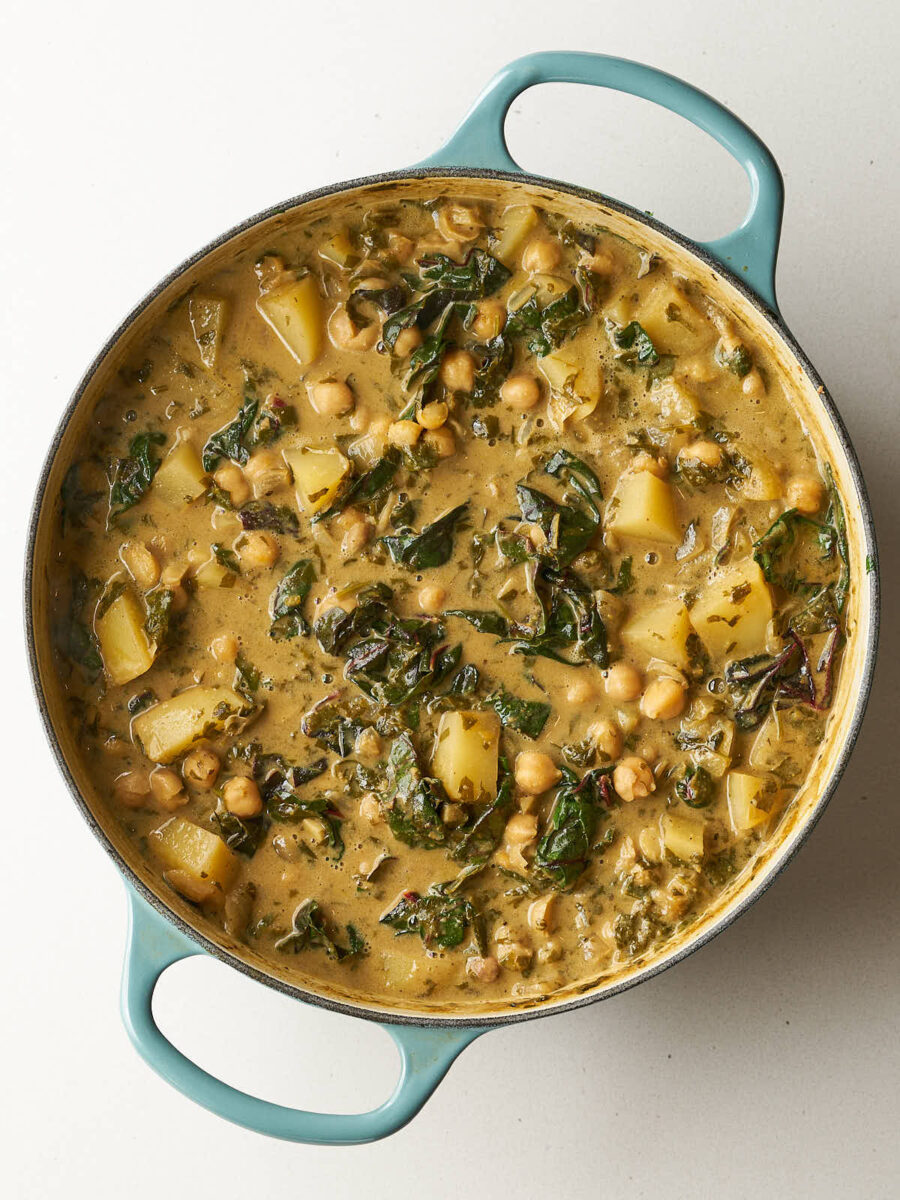 Shawarma chickpea stew with potatoes, chickpeas, and wilted dark green chard simmering in a creamy sauce in a light blue dutch oven.
