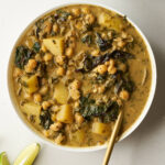 A white bowl of shawarma chickpea stew, garnished with a golden spoon, is placed next to lime wedges. The stew contains chickpeas, potatoes, and dark greens in a creamy, golden sauce.