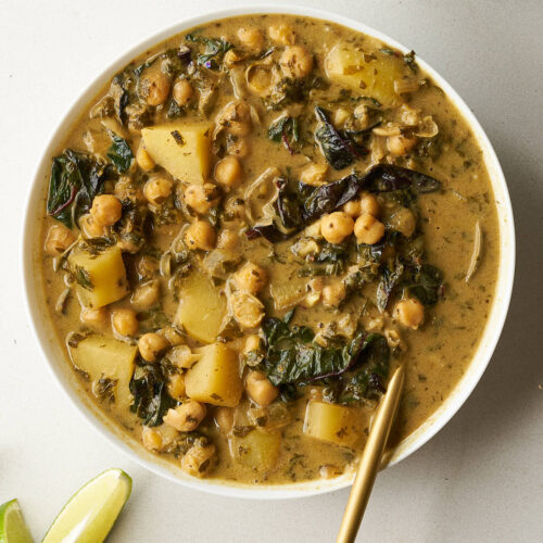 A white bowl of shawarma chickpea stew, garnished with a golden spoon, is placed next to lime wedges. The stew contains chickpeas, potatoes, and dark greens in a creamy, golden sauce.
