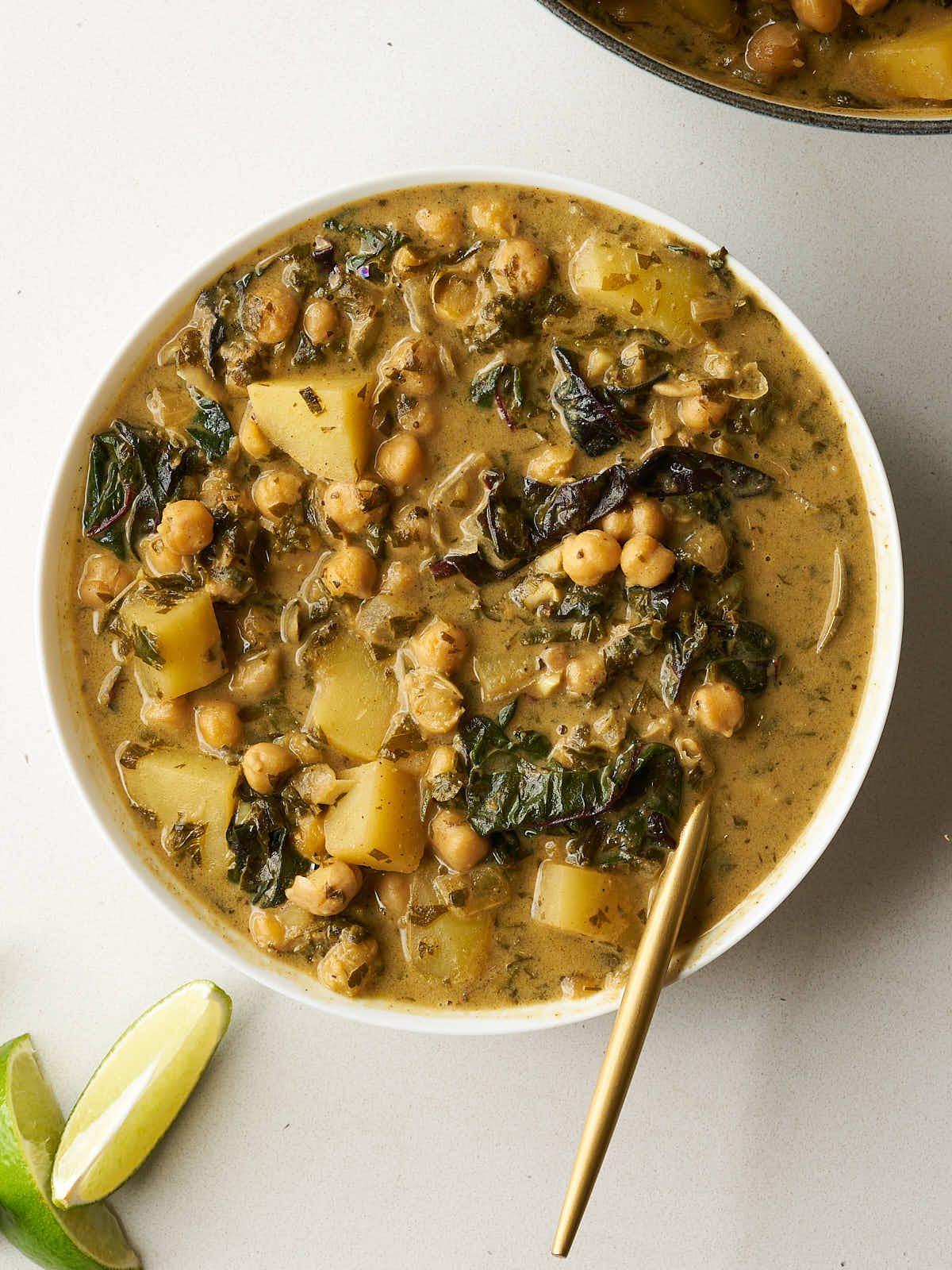 A white bowl of shawarma chickpea stew, garnished with a golden spoon, is placed next to lime wedges. The stew contains chickpeas, potatoes, and dark greens in a creamy, golden sauce.