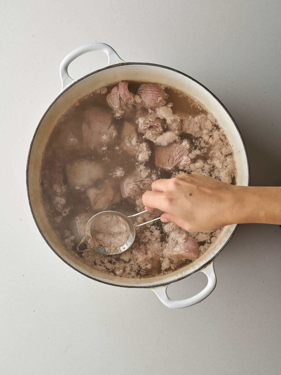 A hand using a small strainer to skim foam and impurities from lamb pieces boiling in a large white enameled pot.
