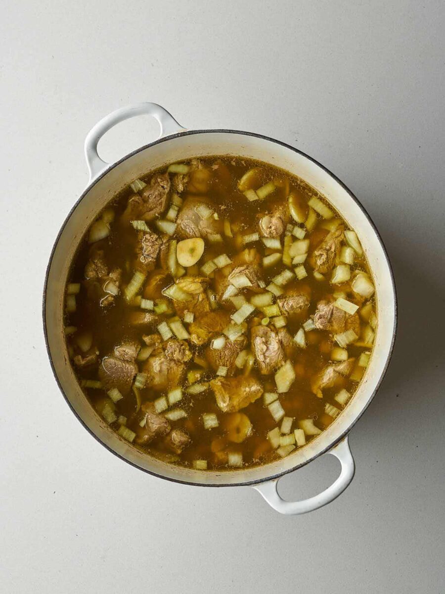 Lamb, diced onion, and slices of garlic submerged in a yellowish broth in a white enameled pot.