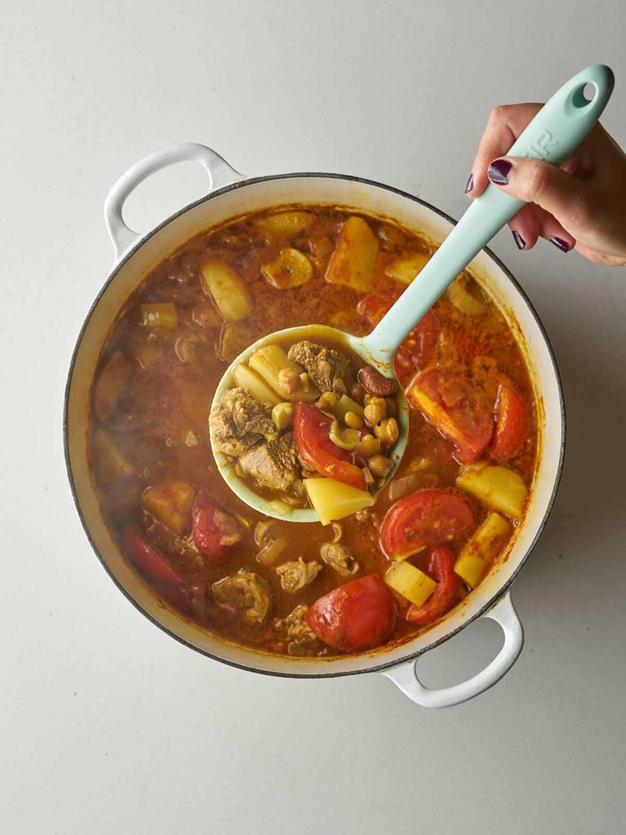 A hand holding a light blue ladle, scooping a serving of the thick stew, showing pieces of lamb, potatoes, beans, and tomato.