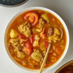 he finished Abgoosht served in a white bowl, showing the rich, red broth and a generous amount of solid ingredients like lamb chunks, beans, and tomatoes, with a gold spoon resting in the bowl.