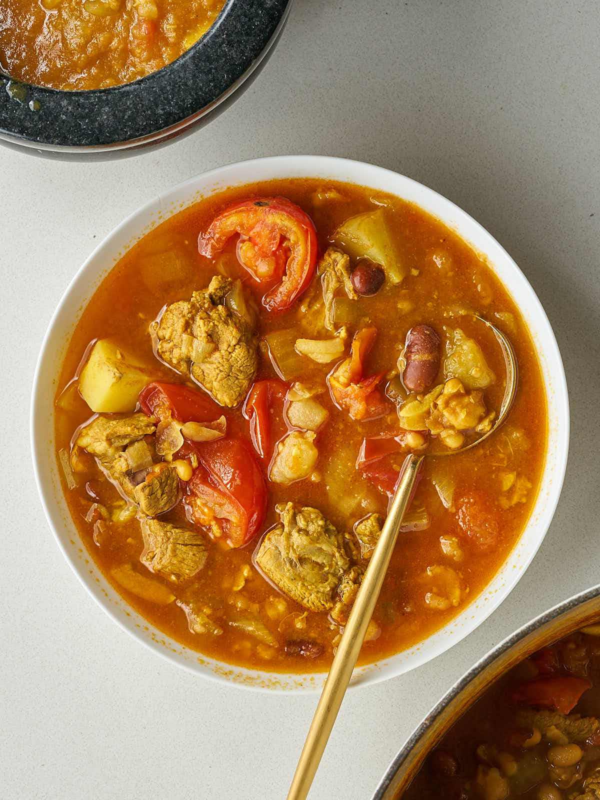 he finished Abgoosht served in a white bowl, showing the rich, red broth and a generous amount of solid ingredients like lamb chunks, beans, and tomatoes, with a gold spoon resting in the bowl.