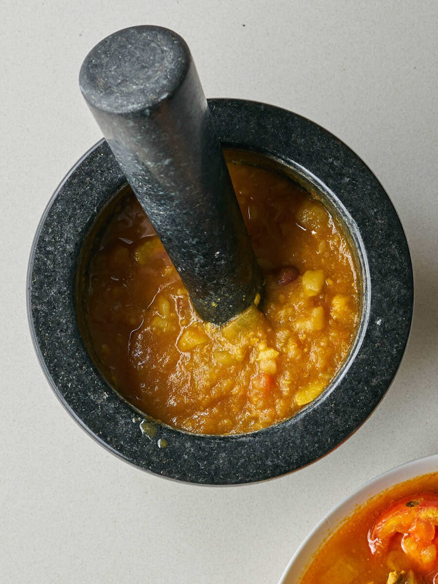 A close-up of a dark stone mortar and pestle being used to mash a portion of the solid ingredients from the stew into a thick paste.