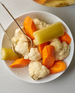 A close-up of a white bowl filled with fermented cauliflower, carrots, and celery pieces, with metal tongs resting on the bowl's edge.