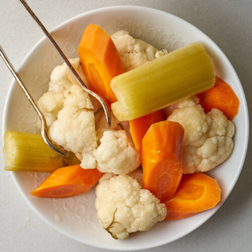 A close-up of a white bowl filled with fermented cauliflower, carrots, and celery pieces, with metal tongs resting on the bowl's edge.