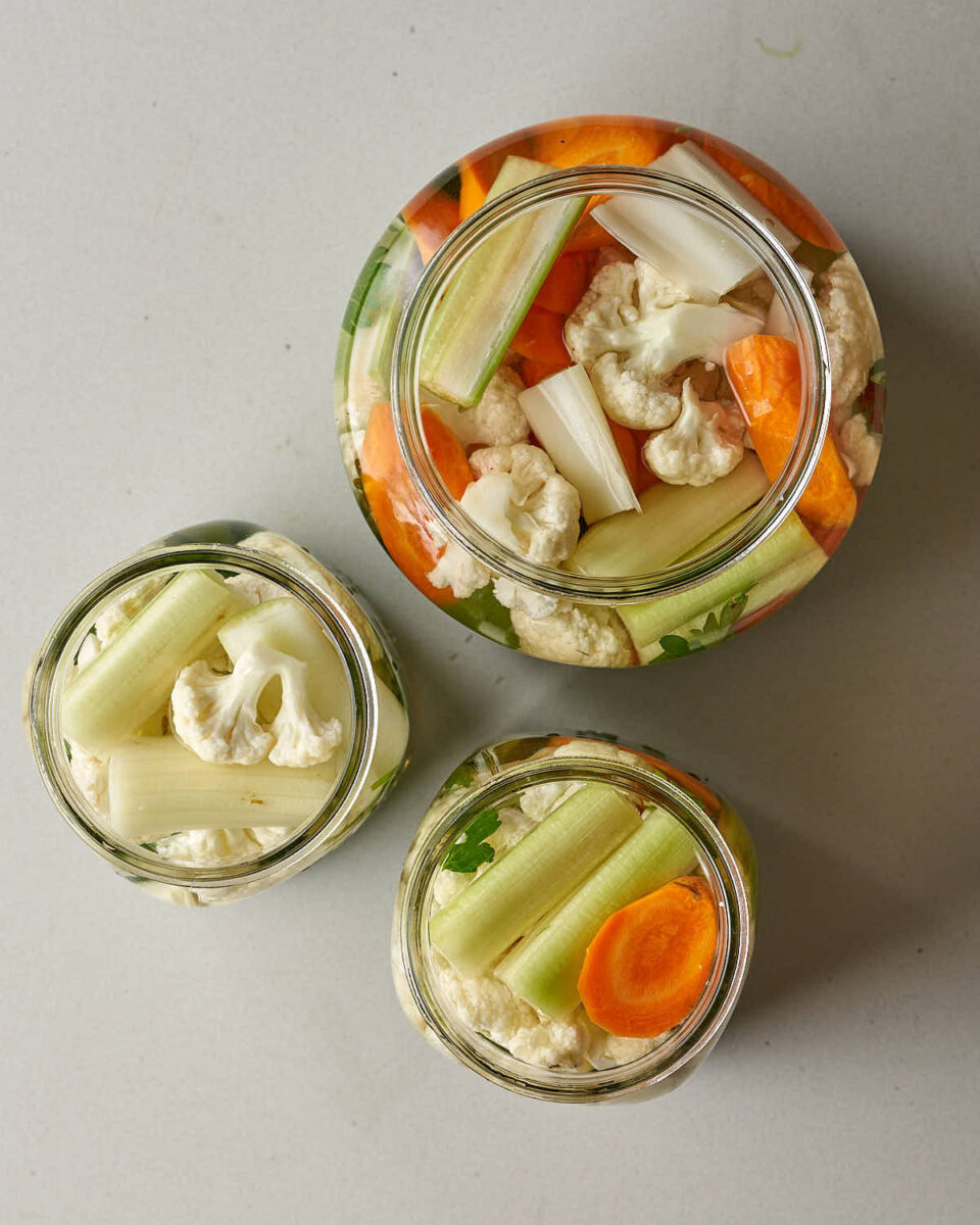 An overhead view of three glass jars filled with chopped cauliflower, carrots, and celery pieces and brine.