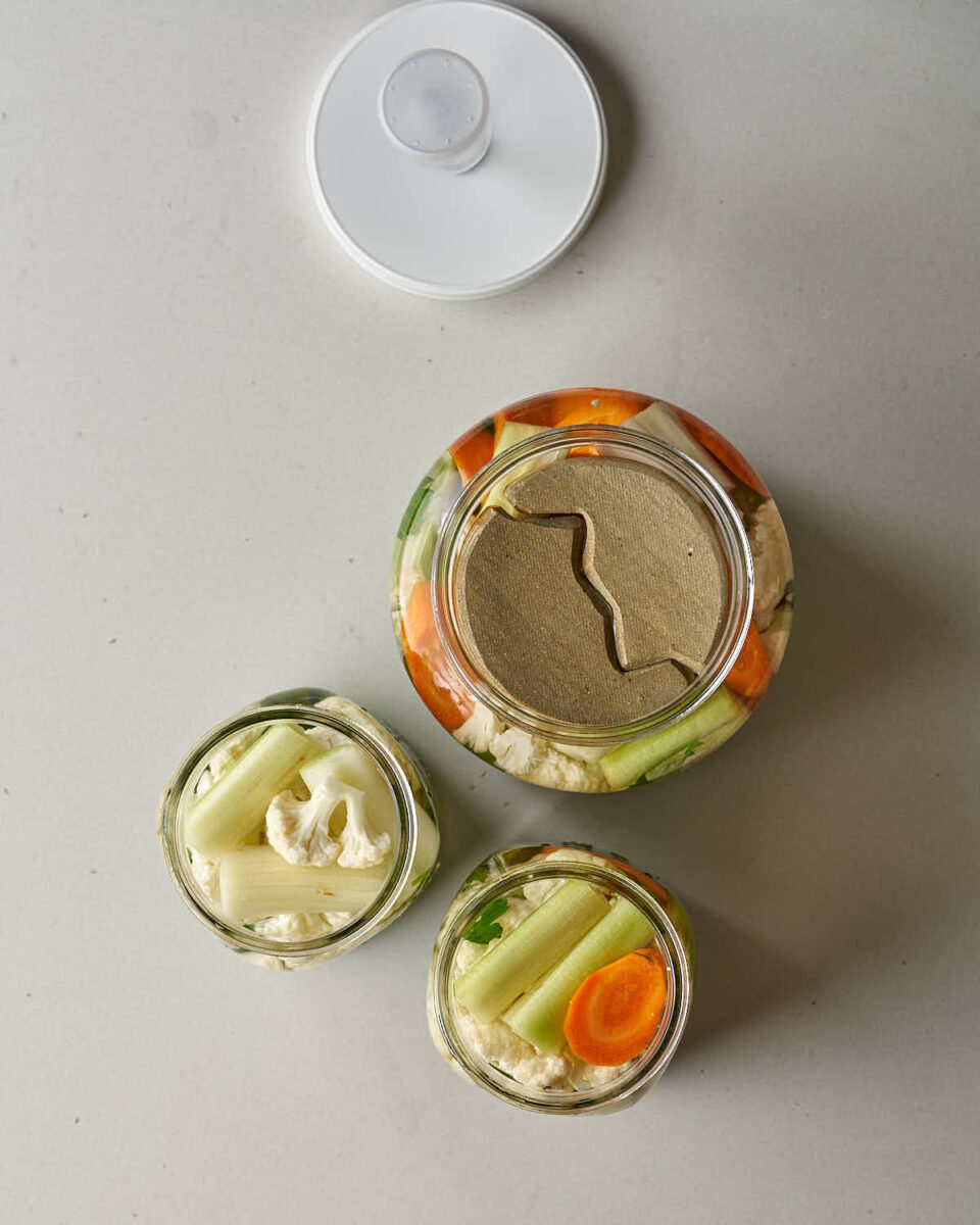 An overhead view of three jars filled with vegetables and brine, one large and two small, with the large jar having a fermentation weight or insert and a white airlock lid lying next to it.