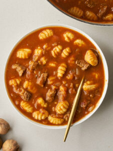 A close-up of a white bowl filled with Gheymeh Gnocchi stew, featuring a gold spoon and a side of dried limes.