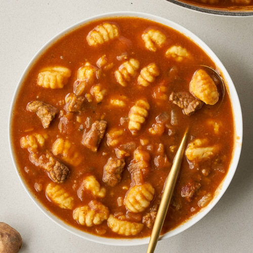 A close-up of a white bowl filled with Gheymeh Gnocchi stew, featuring a gold spoon and a side of dried limes.