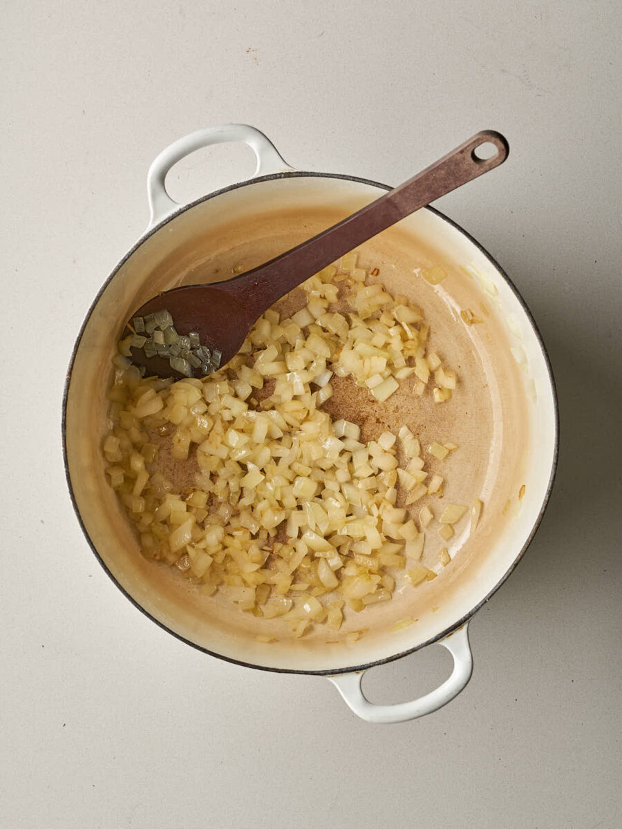 Diced yellow onions being sautéed in a white Dutch oven with a wooden spoon.