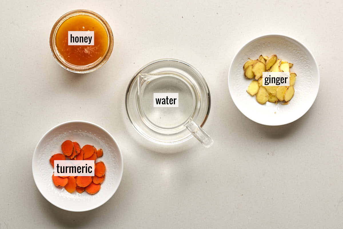 An overhead shot of ingredients in separate white bowls labeled "honey," "ginger," "turmeric," and a central glass of "water."
