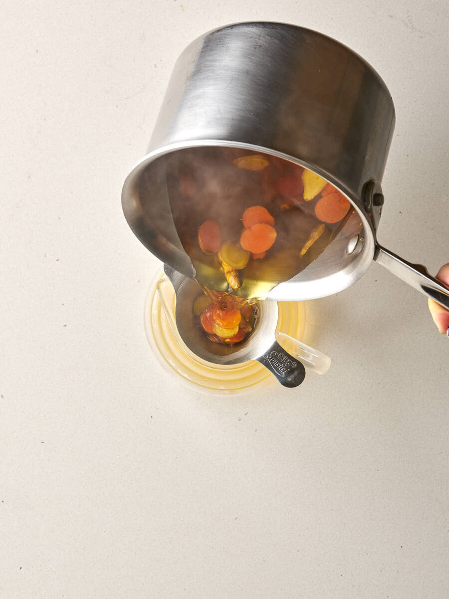 A person pouring hot tea from a stainless steel pot through a metal strainer into a glass carafe, showing the simmered root slices being caught in the mesh.