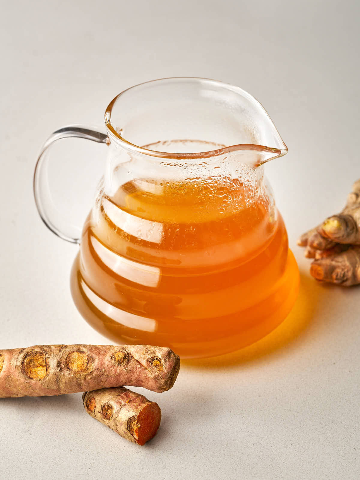 A clear glass carafe filled with amber-colored tea, placed next to whole pieces of fresh turmeric and ginger root.