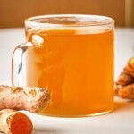 A close-up side view of a glass mug filled with bright orange-gold tea, sitting next to fresh, knobby turmeric roots.