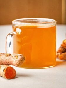 A close-up side view of a glass mug filled with bright orange-gold tea, sitting next to fresh, knobby turmeric roots.