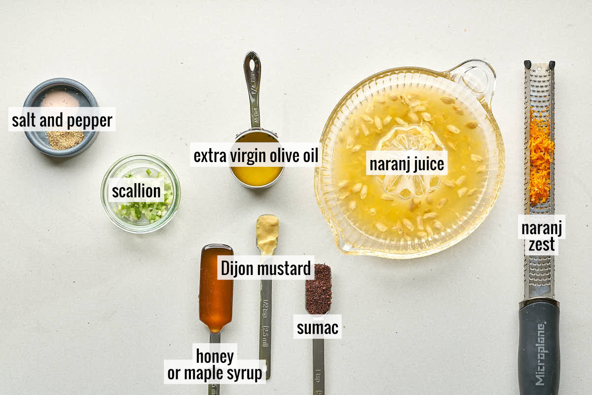 An overhead view of ingredients for Naranj Vinaigrette laid out on a light surface with labels. Ingredients include salt and pepper, chopped scallions, extra virgin olive oil, a bowl of naranj juice, naranj zest on a microplane, Dijon mustard, honey or maple syrup, and sumac.
