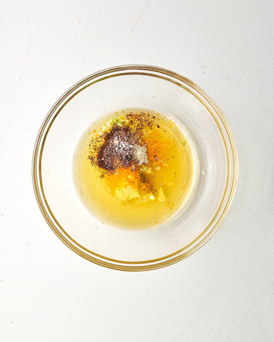 An overhead shot of a clear glass mixing bowl containing the unmixed vinaigrette ingredients, including the juice, oil, spices, and zest centered at the bottom.