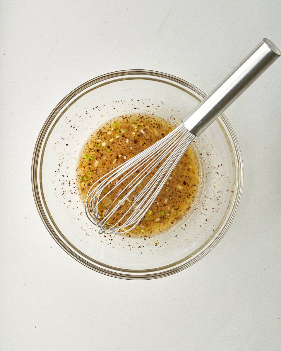 An overhead shot of the glass bowl as the ingredients are being mixed together with a metal whisk. The vinaigrette is becoming emulsified and the spices are dispersed throughout.