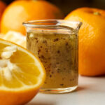 A side-angle close-up of the finished naranj vinaigrette in a small glass pitcher. A sliced bitter orange is in the foreground, with whole oranges blurred in the background.