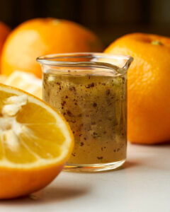 A side-angle close-up of the finished naranj vinaigrette in a small glass pitcher. A sliced bitter orange is in the foreground, with whole oranges blurred in the background.