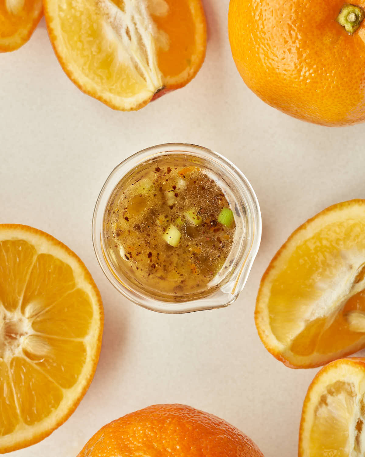 An overhead view of the glass pitcher filled with the finished vinaigrette, surrounded by several whole and halved bitter oranges on a light tabletop.