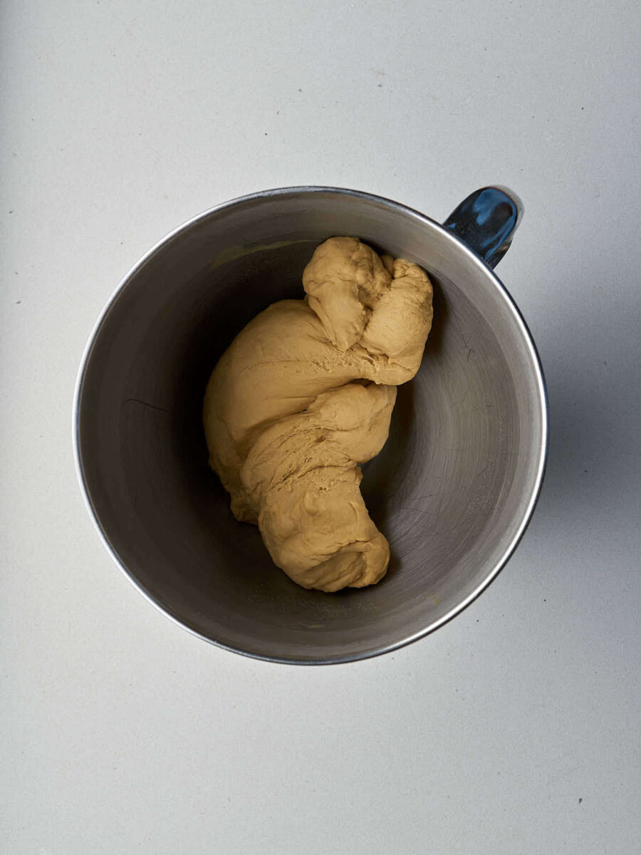 A large, smooth ball of tan-colored bagel dough sits at the bottom of a stainless steel mixing bowl.
