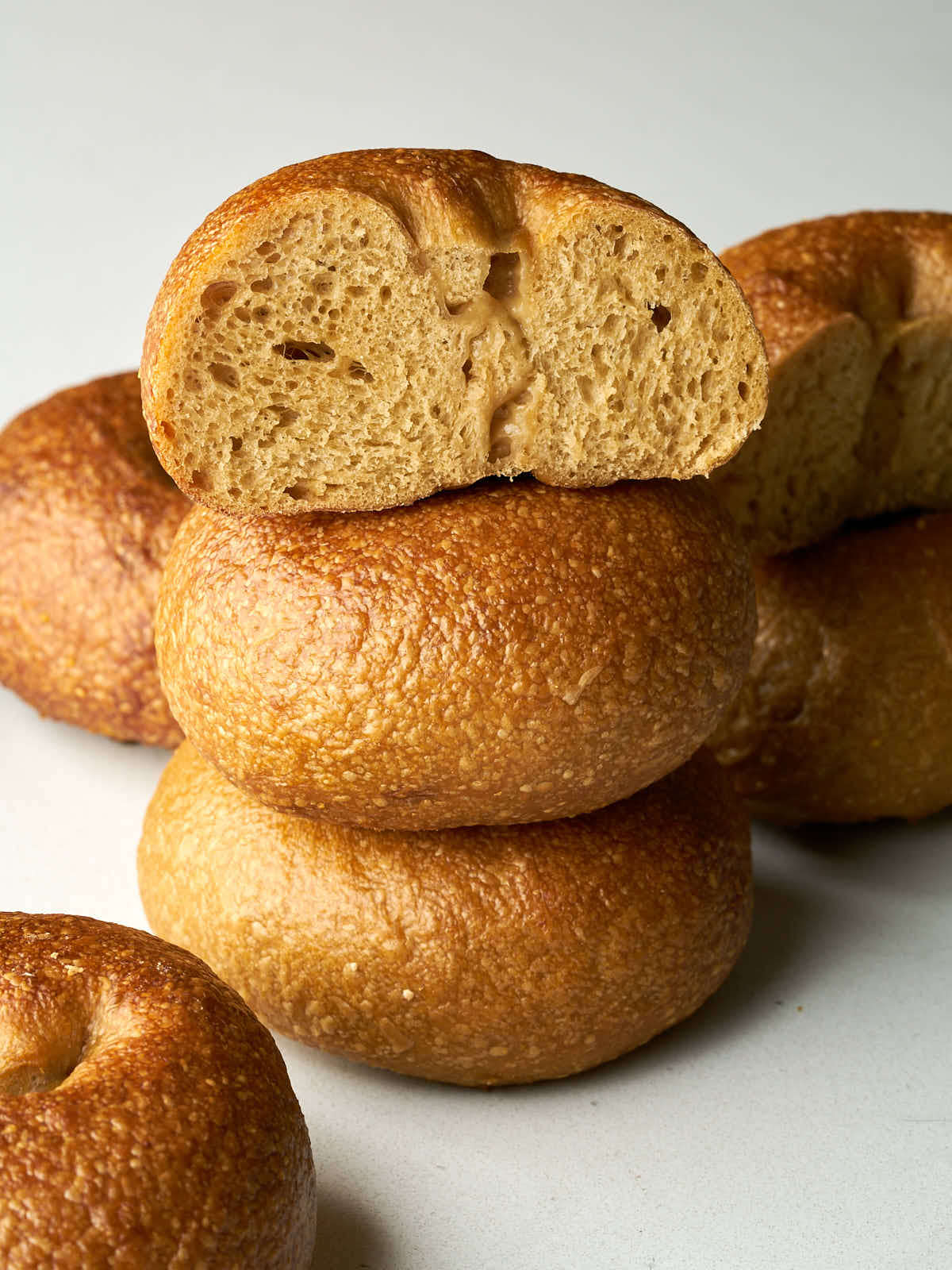 A stack of three golden-brown, baked bagels. The top bagel is sliced in half, revealing a dense, chewy interior.