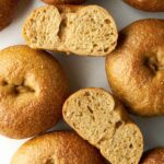 A top-down view of several baked bagels scattered on a white surface, with two sliced in half to show the crumb.
