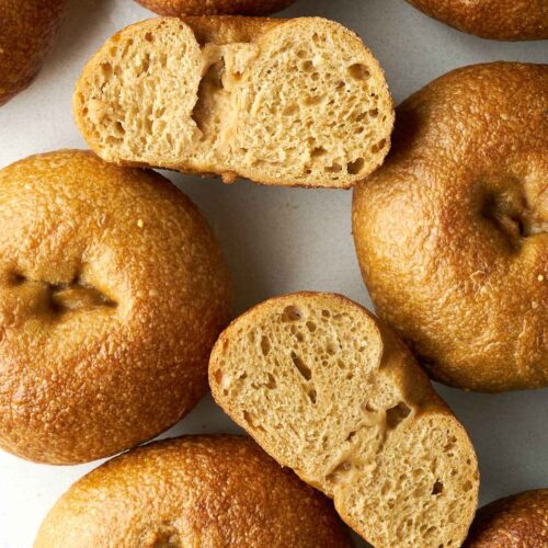 A top-down view of several baked bagels scattered on a white surface, with two sliced in half to show the crumb.