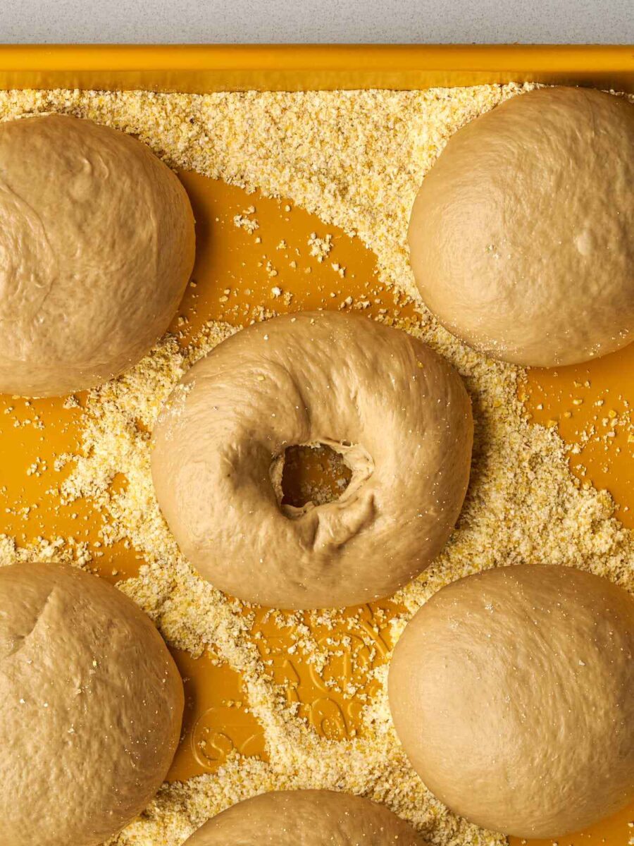 A close-up of the yellow tray where one dough ball has been poked in the center to form a classic bagel ring shape.