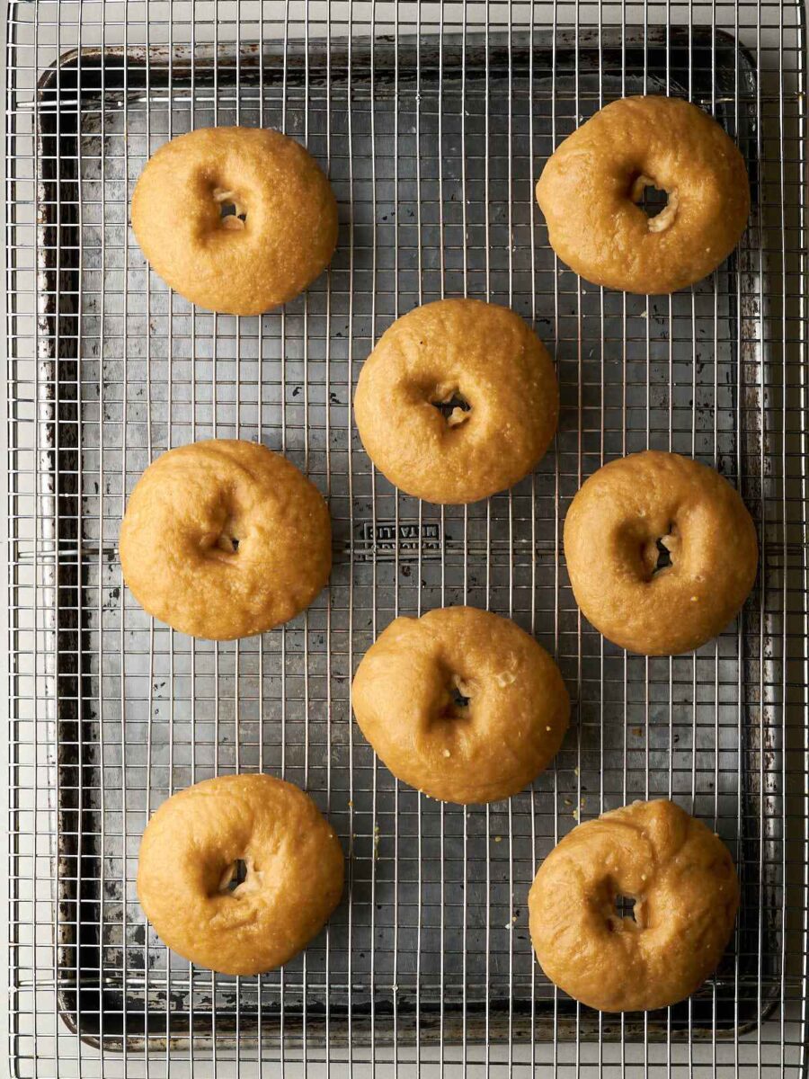 Eight shaped bagel dough rings are arranged on a wire cooling rack set over a metal baking sheet after being boiled. They have a smooth, slightly wet, and pale tan appearance.
