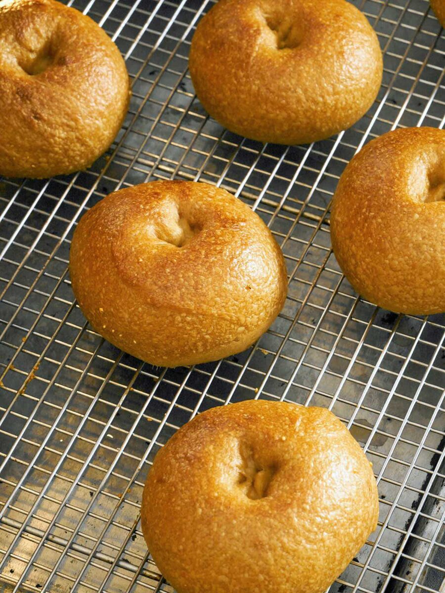 An angled, close-up shot of several bagels on a wire rack, now baked to a glossy, golden-brown color with a textured, bubbly crust.
