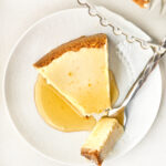 Top-down view of a slice of cheesecake drizzled with orange syrup on a white patterned plate, with a fork taking a small bite.