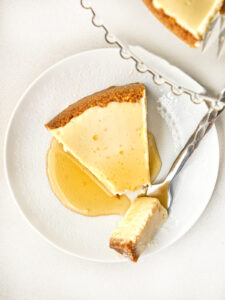 Top-down view of a slice of cheesecake drizzled with orange syrup on a white patterned plate, with a fork taking a small bite.