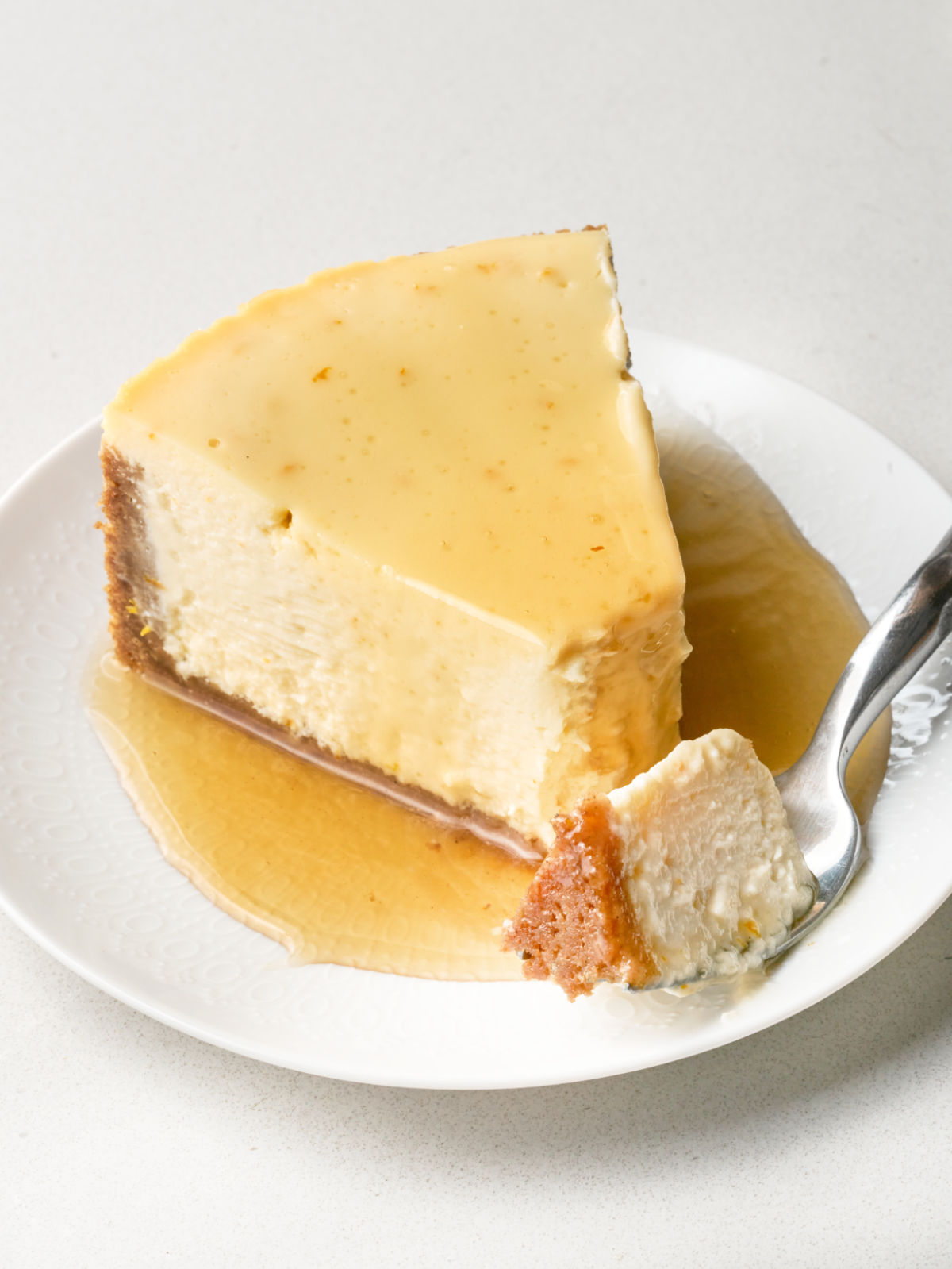 Angled side view of a slice of cheesecake with a graham cracker crust and honey on a plate, with a fork holding a bite in the foreground.
