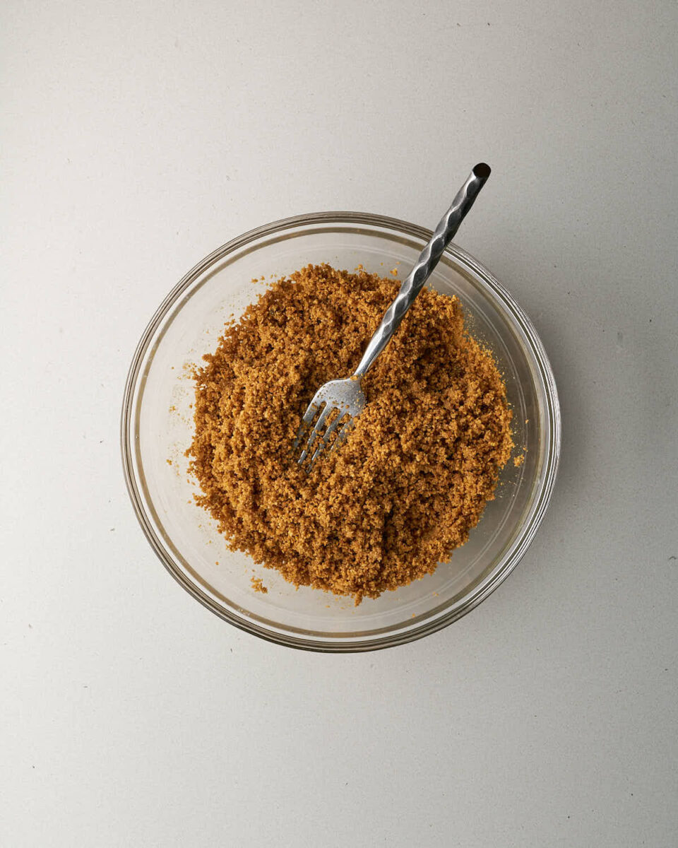 Overhead view of graham cracker crumbs being mixed with a fork inside a clear glass bowl.