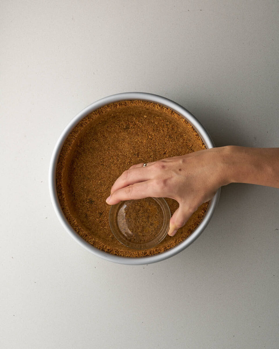 A hand using the bottom of a small glass cup to pack down the graham cracker crust inside a cake pan.