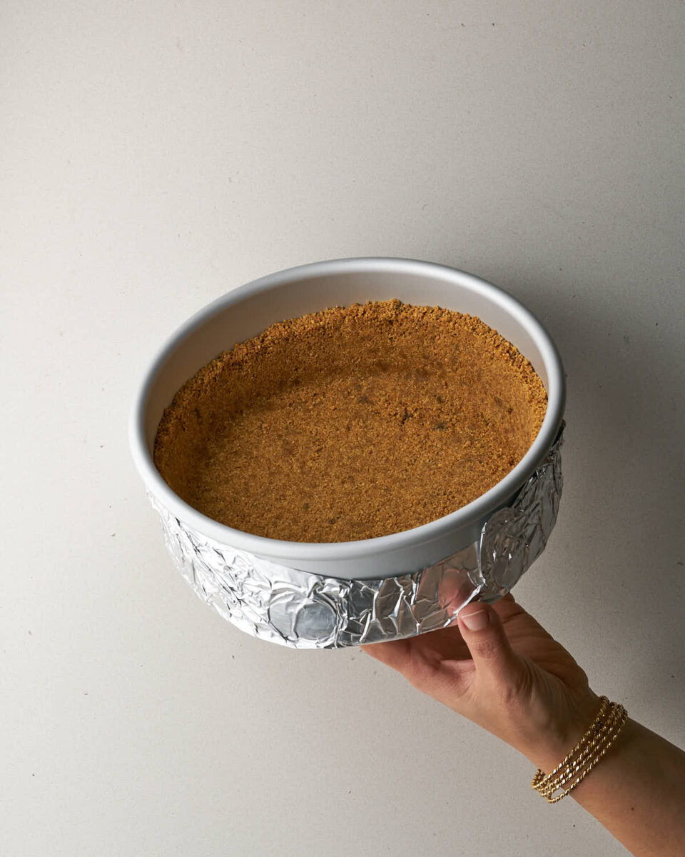 A hand wearing gold bracelets holding a cake pan with a graham cracker crust, wrapped around the bottom and sides with aluminum foil.