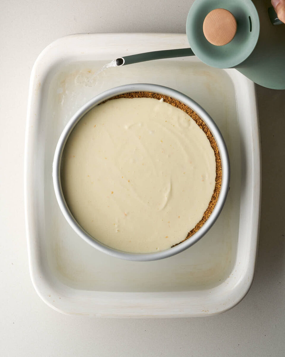 Water being poured from a light green kettle into a large white baking dish surrounding the cheesecake pan to create a water bath.