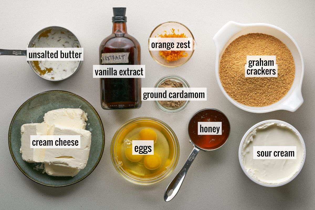 Flat-lay image displaying labeled ingredients for cardamom orange honey cheesecake: unsalted butter, vanilla extract, orange zest, graham crackers, ground cardamom, cream cheese, eggs, honey, and sour cream.