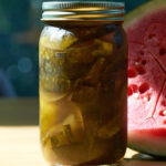Jar of green pickled watermelon rind next to half a watermelon on a wood table.
