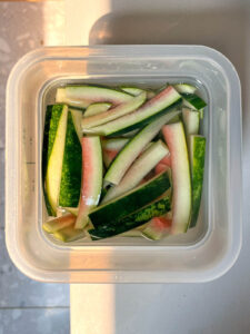 Container of watermelon rinds soaking in a clear brine. The rind strips are white with pale pink edges and dark green skin, viewed from above in a square plastic container under warm sunlight.