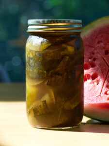 Jar of green pickled watermelon rind next to half a watermelon on a wood table.