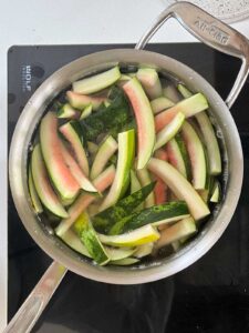 Overhead view of a saucepan filled with watermelon rind pieces soaking in clear brine on an induction cooktop. The rind strips are white with light pink flesh and dark green skin, floating in the liquid as part of the pickling process.
