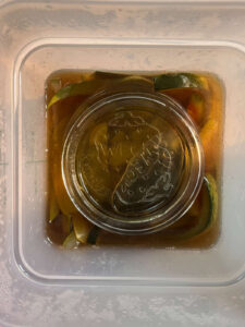 Overhead view of pickled watermelon rinds submerged in amber-colored brine inside a plastic container. A glass jar is placed on top to weigh the rind pieces down and keep them fully submerged during pickling.