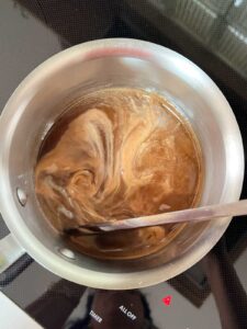 Small saucepan on a stovetop containing amber-colored pickling liquid being stirred with a spoon. Light-colored swirls move through the liquid as ingredients dissolve, viewed from above.
